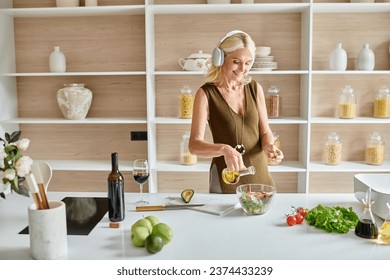 cheerful middle aged woman in wireless headphones making salad near bottle and glass of red wine - Powered by Shutterstock