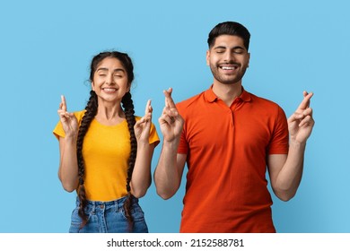 Cheerful Middle Eastern Couple Crossing Fingers For Make A Wish, Smiling Arab Man And Woman Standing With Closed Eyes, Pleading For Fortune And Luck, Posing Together Over Blue Studio Background