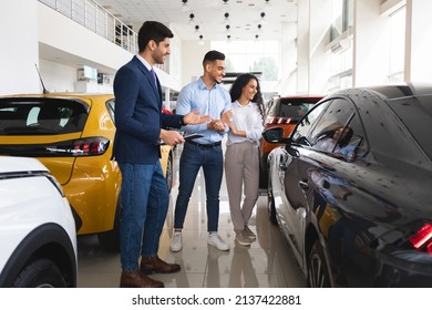 Cheerful Middle Eastern Couple Looking At Black Sports Car, Choosing New Auto At Newest Automobile Dealership, Having Conversation With Male Sales Associate , Full Length Shot, Copy Space