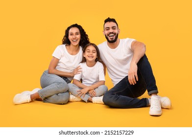 Cheerful Middle Eastern Family Of Three Sitting Smiling To Camera On Yellow Background. Studio Shot Of Arabic Parents And Daughter Hugging Posing Together. Full Length