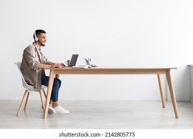 Cheerful Middle Eastern Freelancer Man Working On Pc Online At Home Listening To Music With Wireless Headphones, Making Research For New Project, Sitting At Desk Enjoying Remote Job, Profile Side View