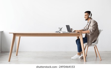 Cheerful Middle Eastern Freelancer Man Working On Pc Online At Home Listening To Music With Wireless Headphones, Making Research For New Project, Sitting At Desk Enjoying Remote Job, Profile Side View - Powered by Shutterstock