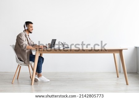 Cheerful Middle Eastern Freelancer Man Working On Pc Online At Home Listening To Music With Wireless Headphones, Making Research For New Project, Sitting At Desk Enjoying Remote Job, Profile Side View