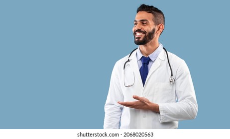 Cheerful Middle Eastern Male Doctor Showing With Hand Free Space Standing Looking Aside Over Blue Background, Wearing White Uniform. Studio Shot. Panorama - Powered by Shutterstock