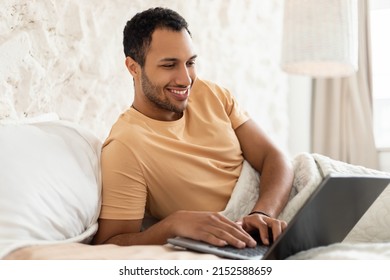 Cheerful Middle Eastern Man Using Laptop Lying In Bed In Bedroom, Working From Home. Male Freelancer Working Online On Computer Browsing Internet And Typing. Freelance, Remote Job