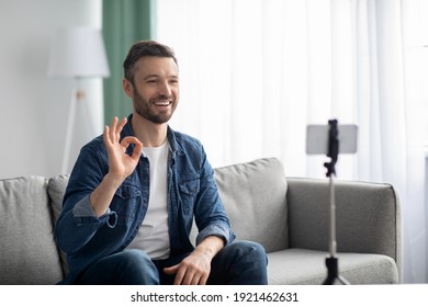 Cheerful Middle-aged Bearded Man Blogger Broadcasting From Home, Using Smartphone And Tripod, Copy Space. Smiling Male Vlogger Waving At Mobile Phone Camera, Shooting Video For Followers