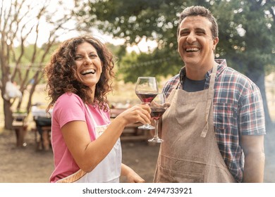 Cheerful middle-aged couple sharing a hearty laugh holding red wine glasses in an outdoor setting. Man and woman in aprons enjoying a carefree moment - joy and connection in leisure activities. - Powered by Shutterstock