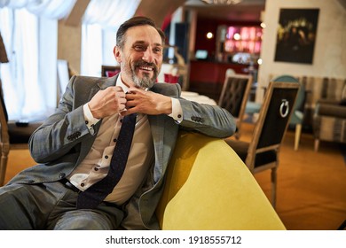 Cheerful middle-aged man smiling while fixing his tie and sitting on a couch