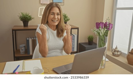 A cheerful middle-aged woman celebrates success at a laptop in a cozy home office setting with plants and decor. - Powered by Shutterstock