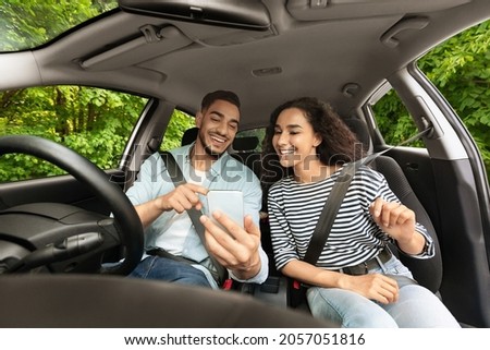 Cheerful middle-eastern guy driver travelling with his beautiful long-haired girlfriend by nice automobile, setting gps on cellphone, couple looking at smartphone screen, shot from car dashboard