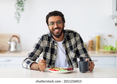 Cheerful Middle-eastern Young Bearded Man In Casual Outfit And Stylish Glasses Enjoying Healthy Lunch At Home, Sitting At Kitchen Table, Eating Vegetable Salad And Drinking Tea, Healthy Deit Concept