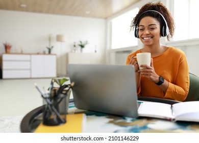 Cheerful Millennial African America Woman Looking At Laptop Screen Drinking Coffee Enjoying Work Or Remote Lecture Sitting At Workplace. Young Female Empolyee Worker Using Computer In Home Office