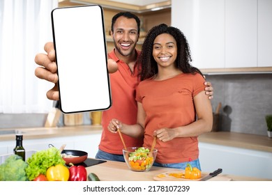 Cheerful Millennial African American Couple Preparing Salad With Organic Vegetables Together Show Big Smartphone With Blank Screen At Kitchen Interior. Cooking At Home, New App, Website, Ad And Offer