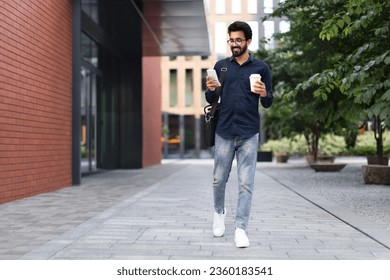 Cheerful millennial arab guy employee going to office in the morning, using phone on the street, drinking takeaway coffee. Happy middle eastern businessman checking email on smartphone, copy space - Powered by Shutterstock