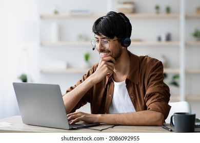 Cheerful Millennial Arabic Man Employee Watching Videos On Internet While Having Coffee Break At Office, Using Modern Laptop And Headset, Looking At Screen And Smiling, Copy Space