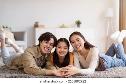 Cheerful Millennial Asian Lady, Guy And Teen Daughter Lie On Floor And Hold Hands In Living Room Interior, Enjoy Spare Time Indoor. Family At Home Together, Relationships During Covid-19 Lockdown