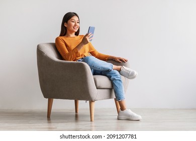 Cheerful millennial Asian woman using mobile phone, chatting on web, working or studying online, sitting in armchair against white wall, copy space. Pretty young lady watching video on smartphone - Shutterstock ID 2091207781