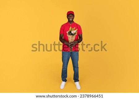 Cheerful millennial black guy courier hold bouquet of flowers, isolated on orange studio background, full length. Delivery of gift at home, holiday holiday, anniversary, Valentine day