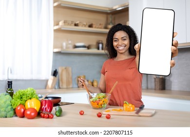 Cheerful Millennial Black Lady Cuts Organic Vegetables At Salad, Shows Smartphone With Blank Screen In Kitchen Interior. App For Food Blog, Social Networks, Household Chores, Cooking Dinner At Home