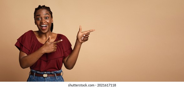 Cheerful millennial black lady posing over colorful background, pointing at copy space and smiling. Beautiful young african american woman showing text or advertisement on beige, panorama