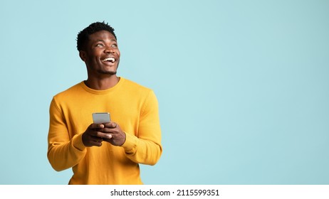 Cheerful millennial black man in casual using modern smartphone, checking social media, looking at copy space for mobile application advertisement and smiling, blue studio background, panorama - Shutterstock ID 2115599351