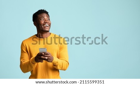 Cheerful millennial black man in casual using modern smartphone, checking social media, looking at copy space for mobile application advertisement and smiling, blue studio background, panorama