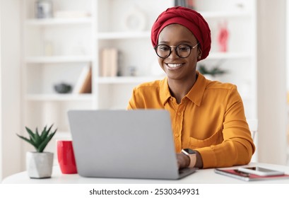 Cheerful millennial black woman entrepreneur working from home, wearing red headscarf and eyeglasses, sitting at workdesk and typing on laptop keyboard, chatting with clients, copy space - Powered by Shutterstock