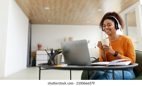 Cheerful millennial black woman watching videos on Internet while having coffee break at home office, using modern laptop and wireless headset, looking at screen and smiling, panorama, free copy space - Powered by Shutterstock