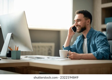 Cheerful Millennial Businessman Relaxing At Workplace In Office With Cellphone And Coffee, Smiling Male Sitting At Desk And Talking On Mobile Phone, Speaking With Family, Having Break At Work - Powered by Shutterstock