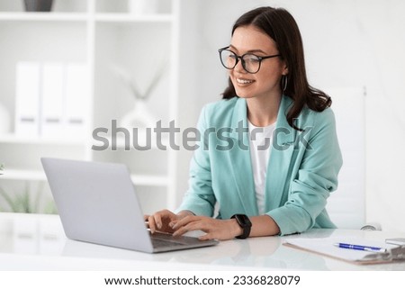 Cheerful millennial caucasian lady in suit and glasses typing on laptop at workplace in office interior. Business with gadget, communication with client remotely, manager work