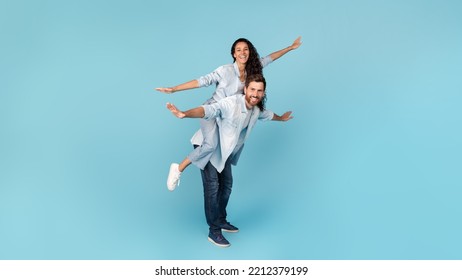Cheerful millennial caucasian man holding middle eastern wife on his back, doing plane, have fun, isolated on blue background, studio. Ad and offer, love and relationship, couple enjoy freedom, love - Shutterstock ID 2212379199