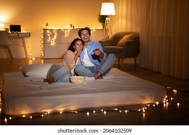 Cheerful Millennial Couple Watching Television Enjoying Movie And Eating Popcorn At Home In The Evening. Spouses Relaxing On Weekend Night Together. Low Light - Powered by Shutterstock