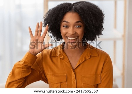 Cheerful millennial curly african american lady waving hand, has video call in living room interior, close up. Meeting at home, communication remotely headshot and hi gesture