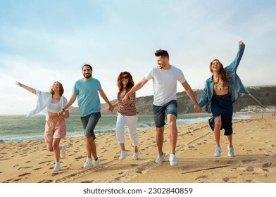 Cheerful millennial european and arab friends have fun, enjoy free time, jump and run on ocean beach, summer party, full length. Walk, vacation together, holiday and trip outdoor