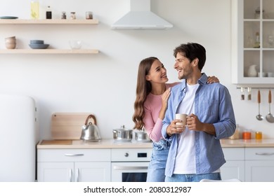 Cheerful millennial european wife hugs husband with cup of hot drink in minimalist kitchen interior. Covid-19 pandemic, self-isolation, couple enjoy coffee together at home, breakfast in morning - Powered by Shutterstock