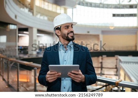 Cheerful millennial islamic guy engineer with beard in hard hat, suit checks project on tablet, looks at free space in factory office interior. Modern tech, professional work in industrial production