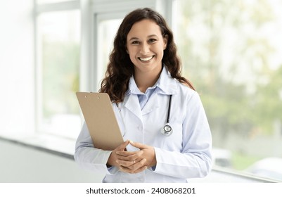 Cheerful millennial lady doctor in white coat, with stethoscope and clipboard standing next to window at clinic, hospital, copy space. Medical exam, health care consultation, prescription