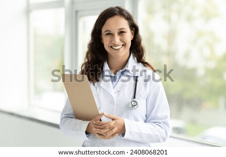 Cheerful millennial lady doctor in white coat, with stethoscope and clipboard standing next to window at clinic, hospital, copy space. Medical exam, health care consultation, prescription