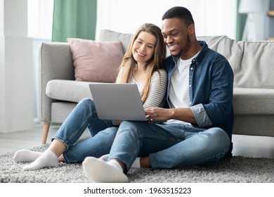 Cheerful Millennial Mixed Couple Spending Time With Laptop At Home, Happy Young Interracial Lovers Sitting On Floor With Computer, Watching Movies Or Shopping Online Together, Free Space