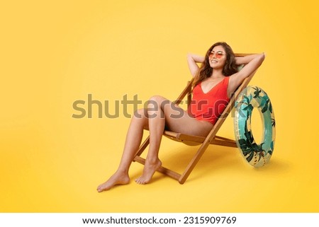 Cheerful millennial mixed race lady in swimsuit and sunglasses with inflatable ring sit in deck chair, enjoy vacation, isolated on yellow studio background. Holiday in summer, weekend and free time