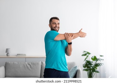 Cheerful millennial muscular attractive european man athlete do arm exercises, warm up in living room interior, empty space. Sport at home, workout and body care, new normal during covid-19 outbreak