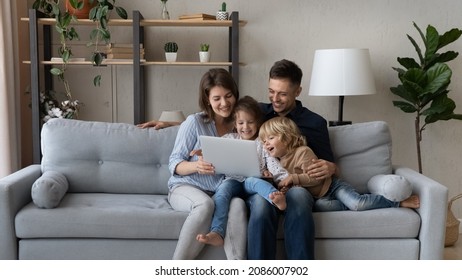 Cheerful Millennial Parents Resting With Gen Z Kids On Couch, Making Video Call, Watching Movie Online, Internet TV On Laptop Computer, Having Fun, Laughing. Happy Family Enjoying Weekend