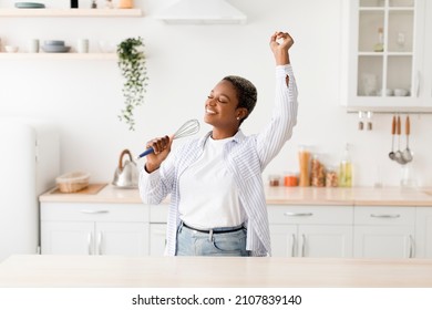 Cheerful millennial pretty black female singing at appliance as imaginary microphone, dancing in scandinavian kitchen interior alone. Fun at home alone, lady superstar enjoy free time, copy space