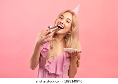 Cheerful millennial woman eating tasty cake, wearing birthday hat, celebrating special occasion on pink studio background. Gorgeous blonde lady enjoying yummy dessert on B-day party