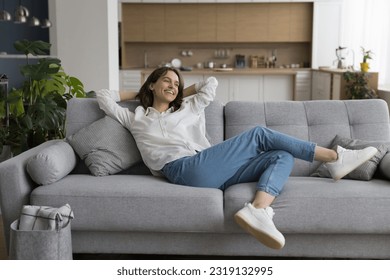 Cheerful millennial woman relax on sofa in studio apartment, leaned on soft comfortable couch with hands behind head, smile enjoy carefree leisure at smart home with climate control for comfort life - Powered by Shutterstock