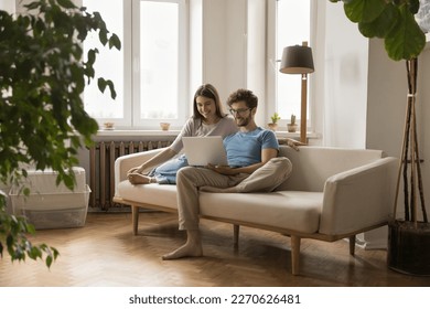 Cheerful millennial young husband and wife using laptop for online communication together, resting on couch, holding computer, talking on video call, watching TV, movie on Internet - Powered by Shutterstock