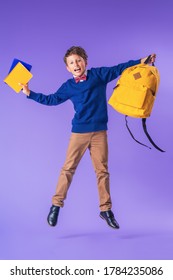 Cheerful Mischievous Schoolboy In Uniform With Backpack And Books Jumps On Purple Background. Dynamic Images That Go Back To Concept School. Beginning Holidays. Back To School. Boy Is Ready To Study.