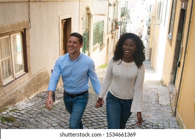 Cheerful mix raced couple enjoying vacation outside. Young man and woman walking up old town street, holding hands and smiling. Happy date concept - Powered by Shutterstock