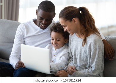 Cheerful Mixed Race Family Sitting Together On Comfortable Sofa In Living Room, Holding Laptop On Knees, Looking At Screen, Playing Online Game, Watching Movie Or Cartoons, Shopping In Internet Store.