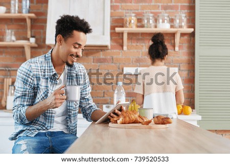 Cheerful mixed race male watches comedy on tablet, uses free internet connection, drinks coffee and eats delicious croissants. Woman stands at cooker, prepares breakfast for family in background